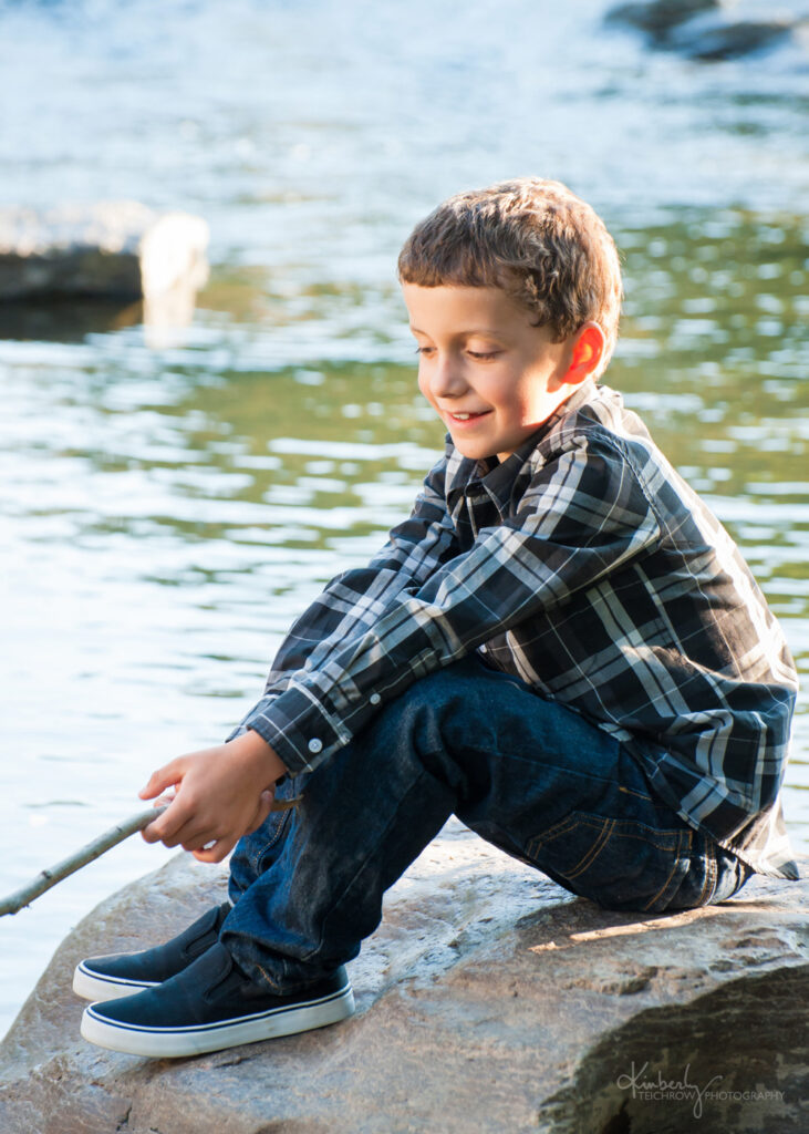 Young boy siting by the bank of a water body playing a stick